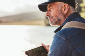 Close up portrait of mature man wearing cap sitting at a lake and looking at a view.